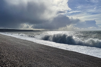 Big waves crashing up onto Chesil Beach, under cloudy, overcast skies.