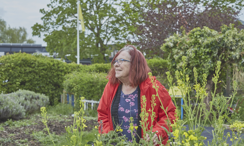 A woman stands by a vegetable patch in a community garden, surrounded by greenery, holding a bowl of freshly collected vegetables.