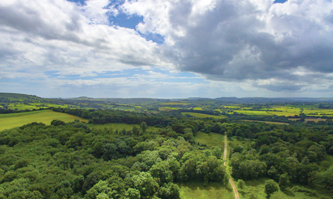 A landscape shot of Powerstock Common taken from above. Green countryside can be seen, with sun behind clouds in the blue sky. 